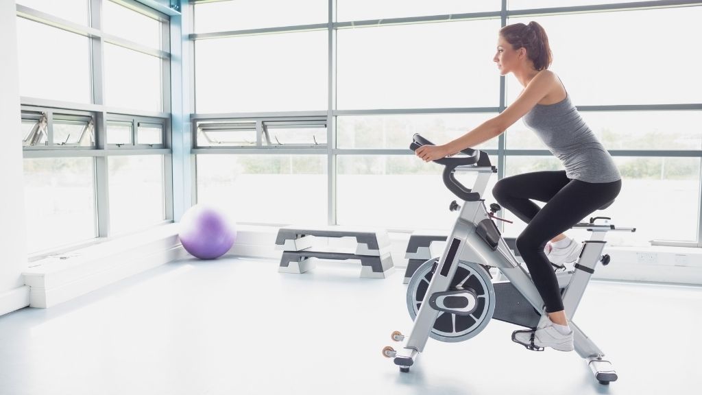 Träningsklädd kvinna sitter på en motionscykel i stort, ljust rum med träningsredskap runtomkring