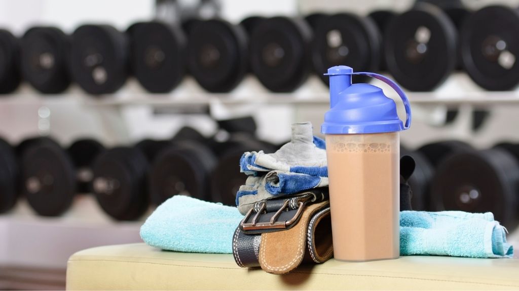 Bilden föreställer en bänk som har flera träningsrelaterade saker på sig. På bänken finns en handduk, ett par träningshandskar, ett tyngdlyftarbälte samt en proteinshake. I bakgrunden syns flera hantlar som ligger i ett ställ.