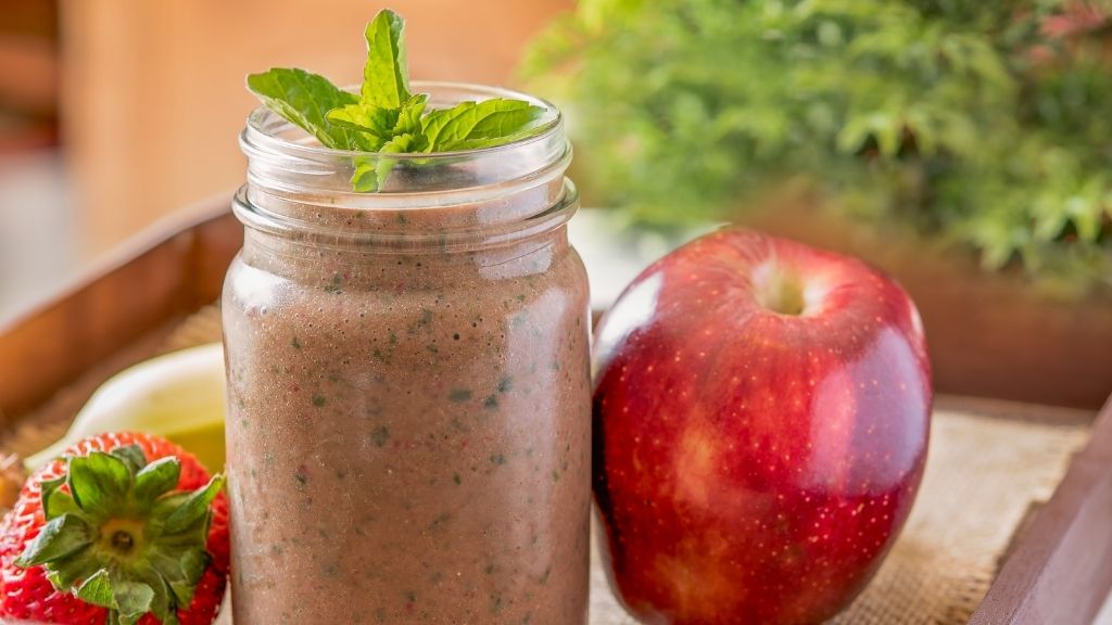 Chokladshake toppad med myntablad i ett glas med ett rött äpple och en jordgubbe bredvid